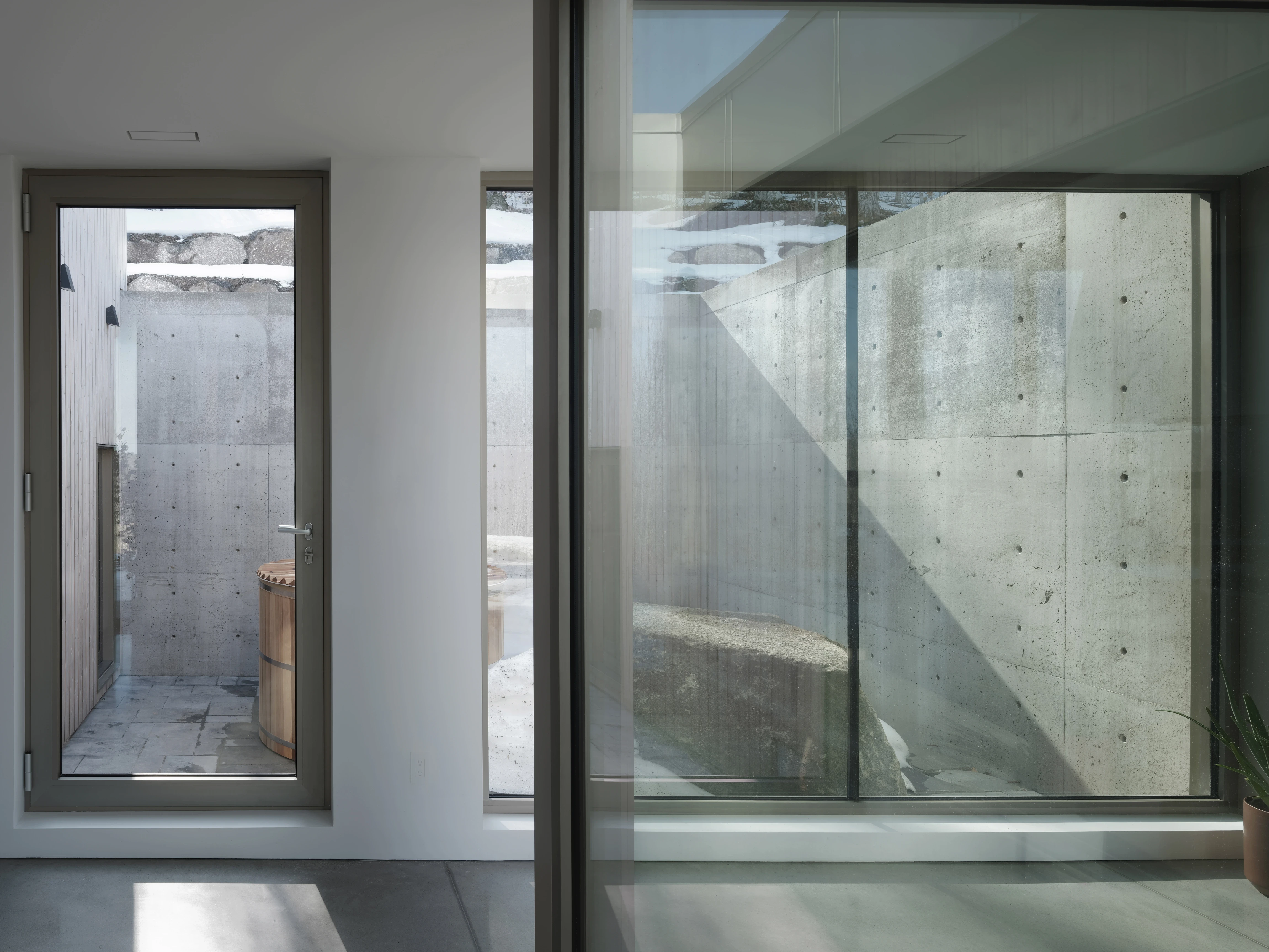 Vue intérieure d’un espace résidentiel minimaliste, murs en béton brut, grandes baies vitrées laissant entrer la lumière naturelle et ouvrant sur une cour extérieure.
