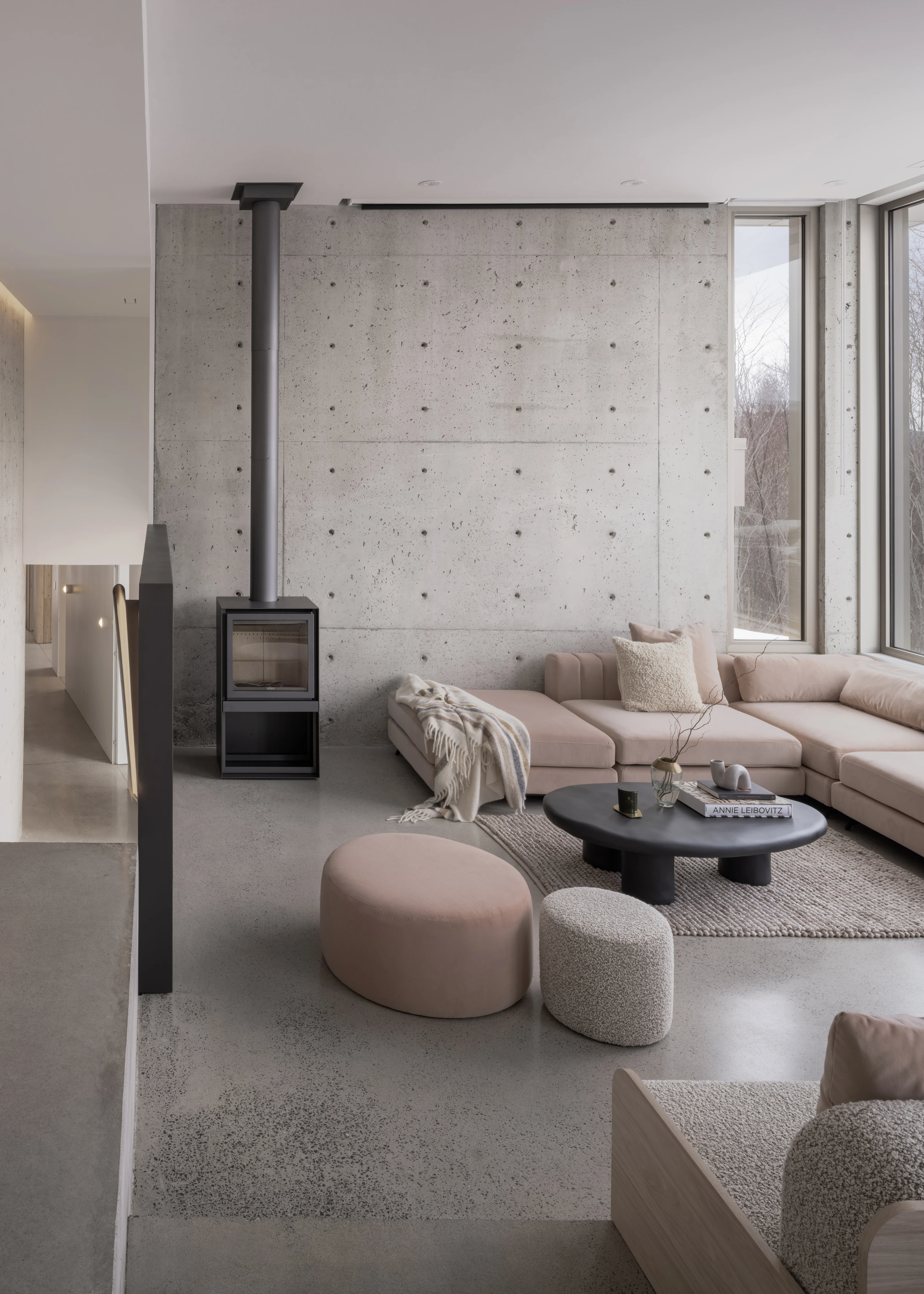 Salon contemporain avec murs en béton brut, foyer au bois, mobilier aux tons neutres et grande fenestration sur le paysage.