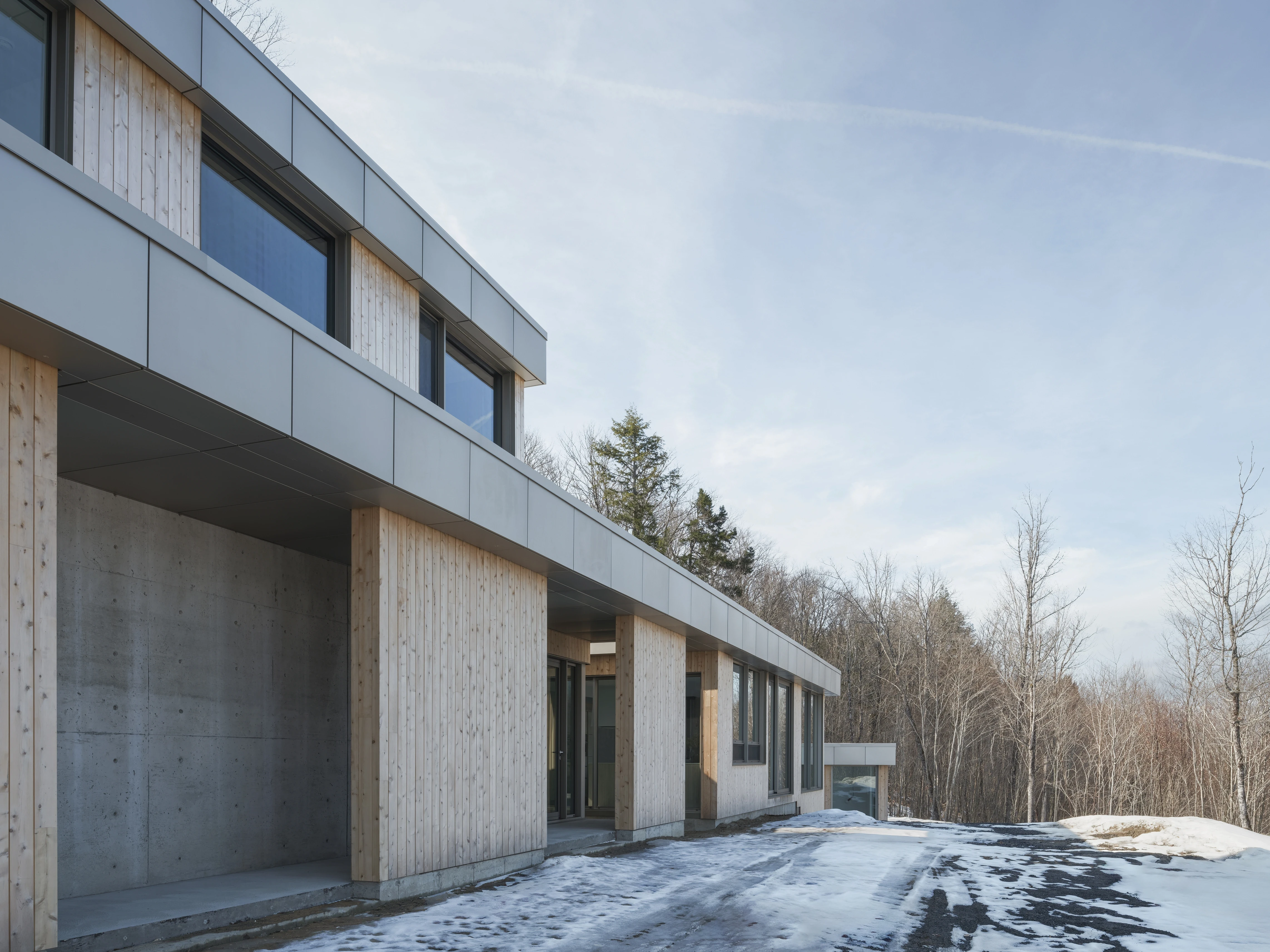 Façade extérieure d’une résidence contemporaine en béton et bois, volumes horizontaux et grandes ouvertures, intégrée à un paysage forestier hivernal au Québec.