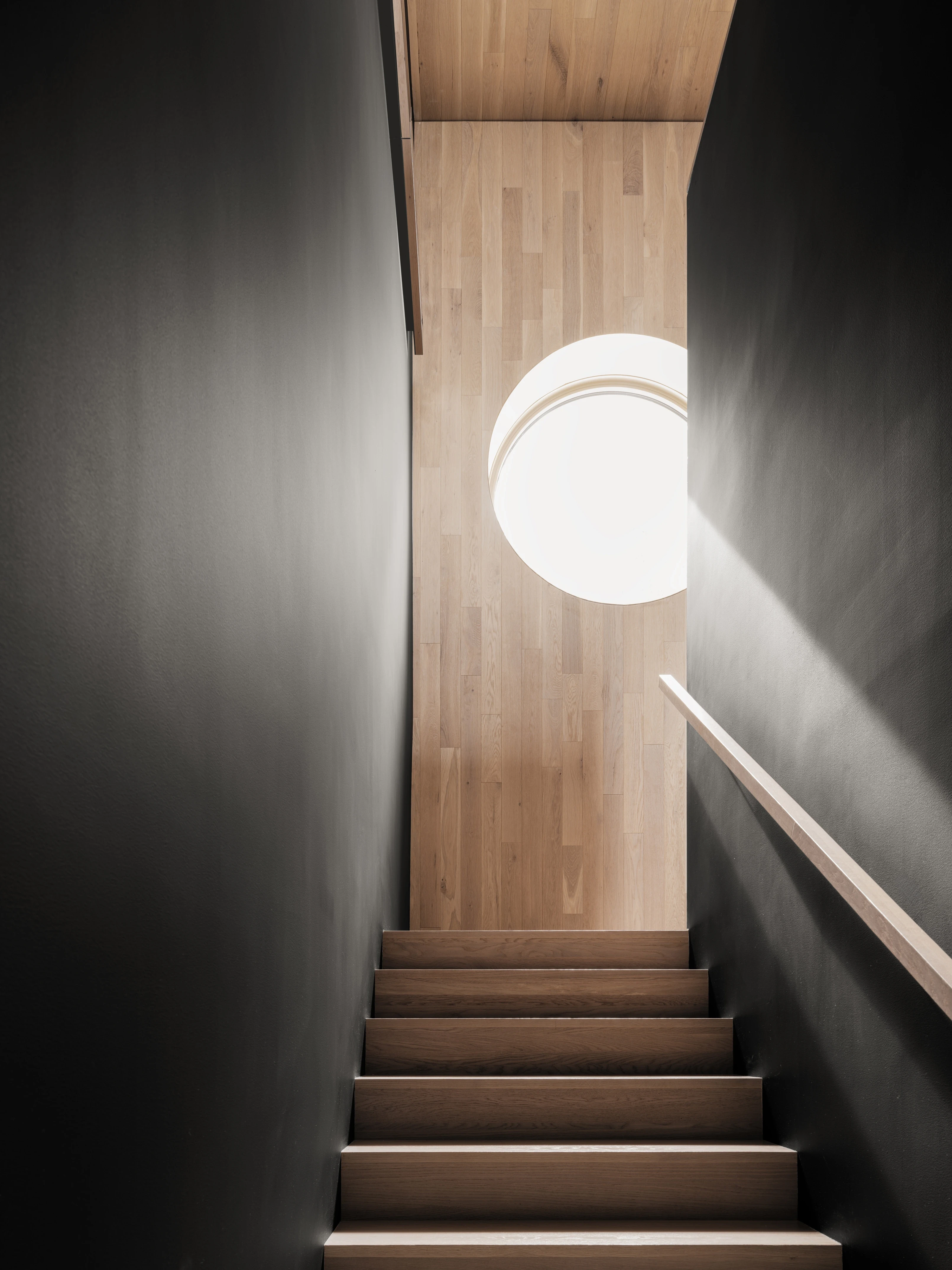 Escalier intérieur contemporain en bois, encadré de murs sombres, éclairé par un puits de lumière circulaire apportant une lumière naturelle zénithale.